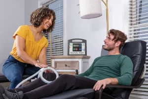 A man sitting in a reclined chair with a woman next to him applying PEMF therapy to his knee, using the Butterfly Loop PEMF attachment from MagnaWavePEMF.